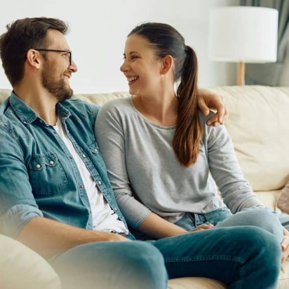 A cheerful couple sits closely on a beige couch, smiling at each other."