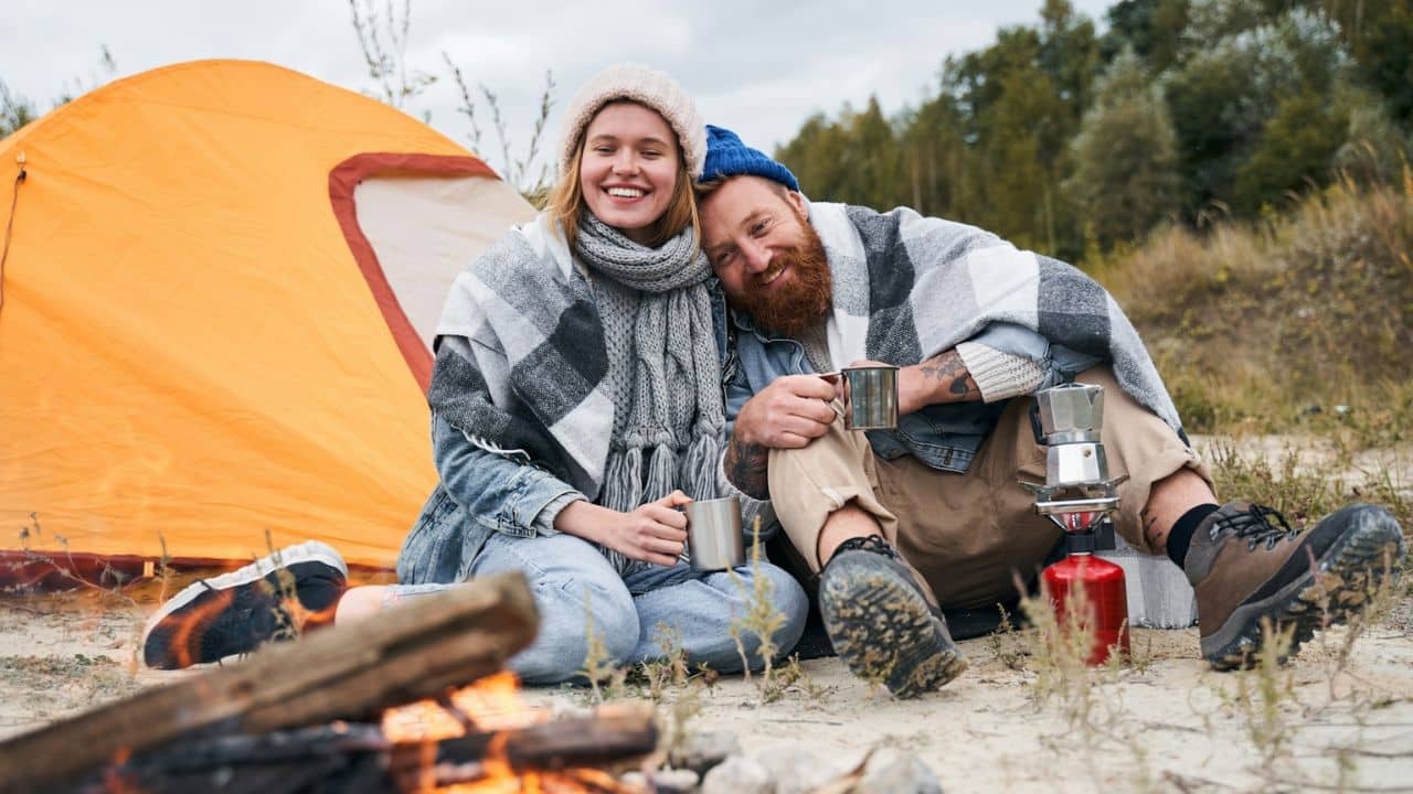 A happy couple sits by a campfire, wrapped in blankets beside a tent.