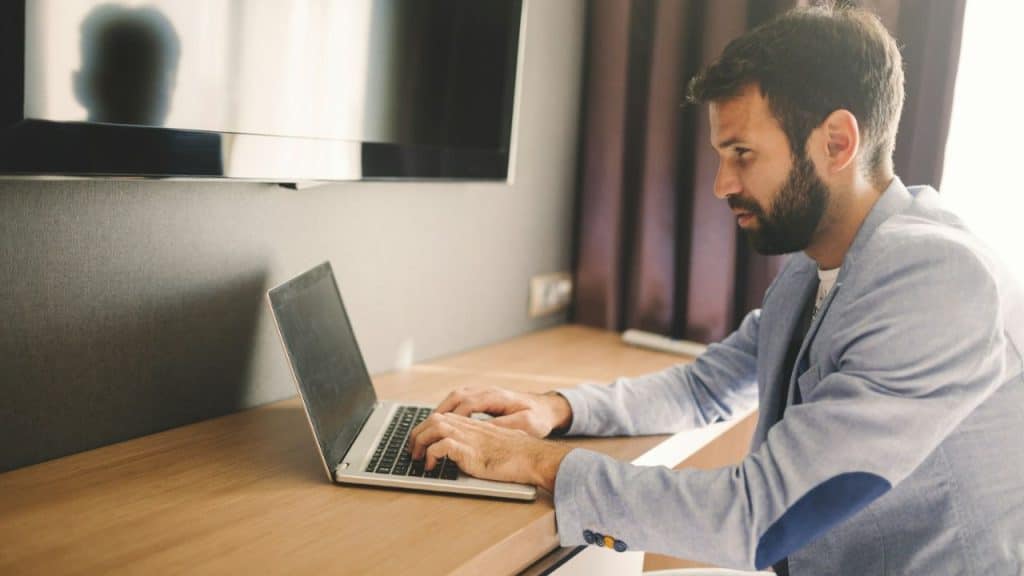 A bearded man in a light blue jacket intently types on a laptop.