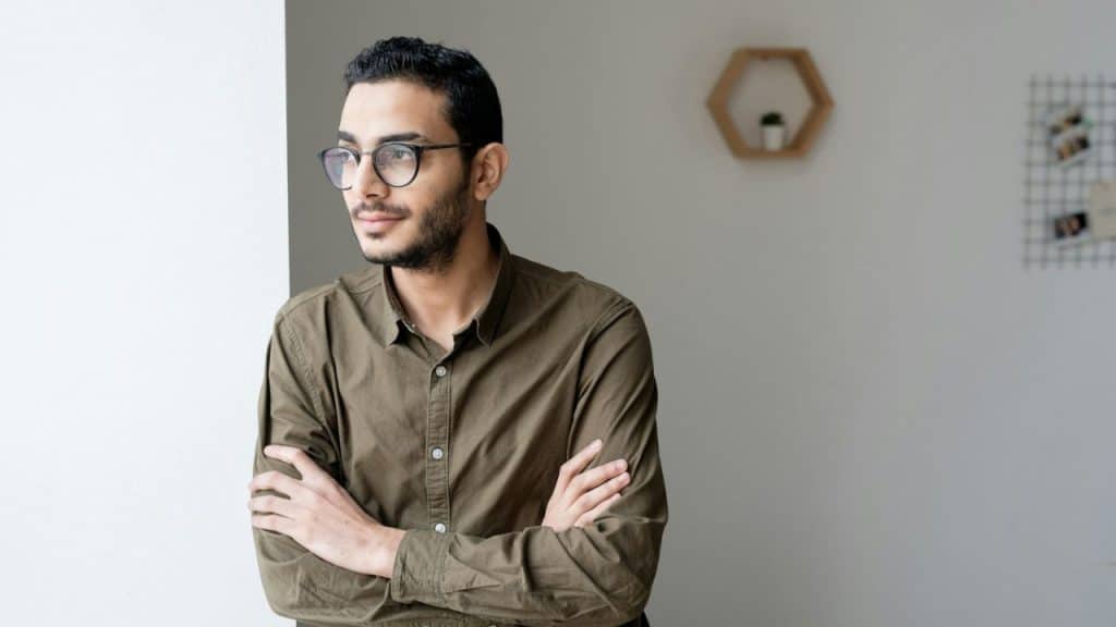 A man with glasses and a beard stands with crossed arms, looking out a window.