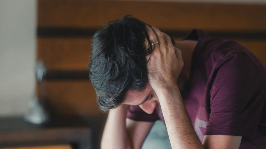 A man with dark hair sits with his hands on his head, looking distressed.
