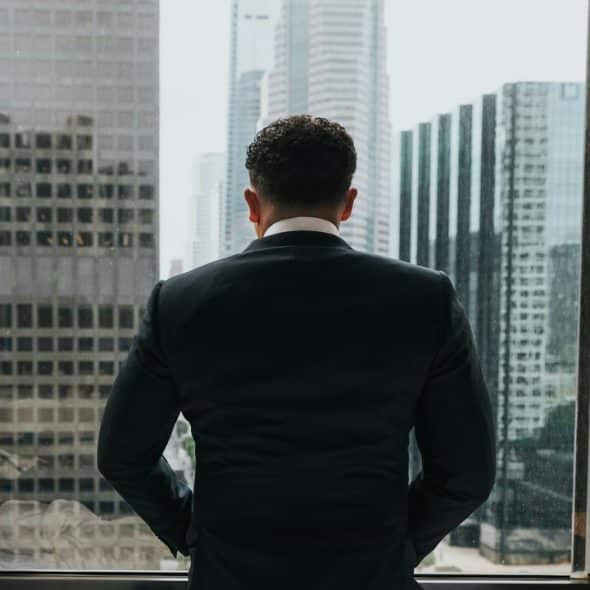 A businessman in a suit standing by a window overlooking city skyscrapers.