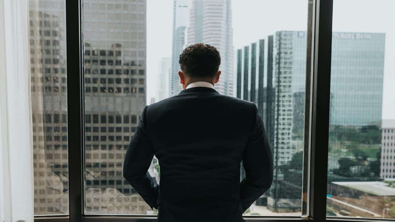 A businessman in a suit standing by a window overlooking city skyscrapers.