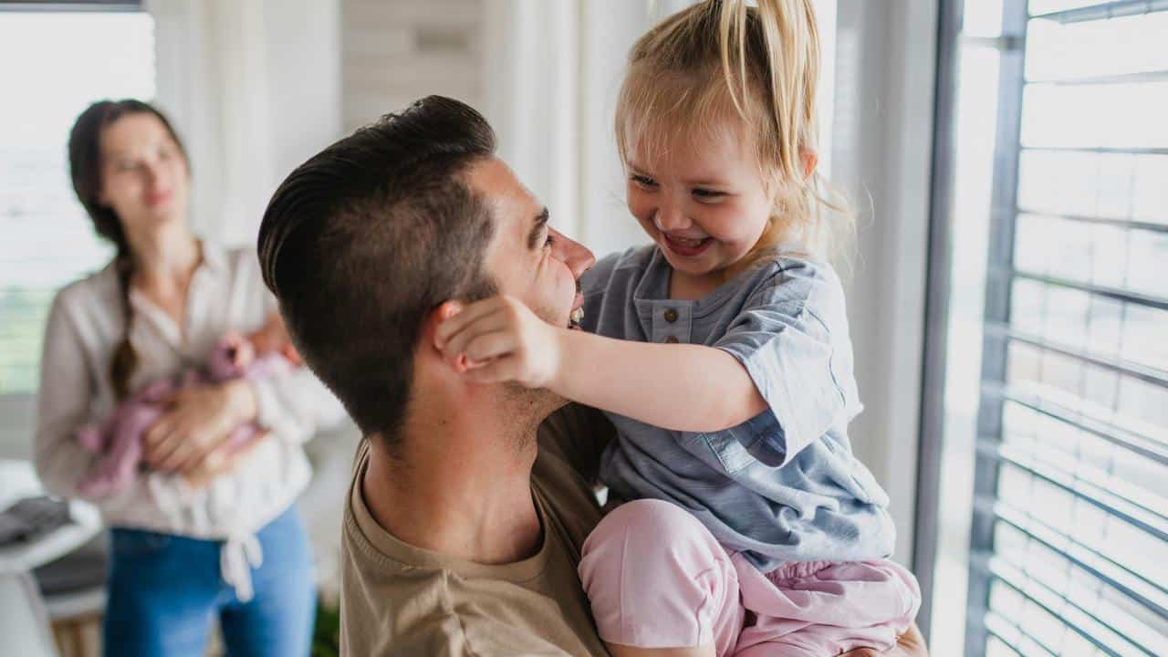 A father holding his daughter while the mother holds a baby in the background.