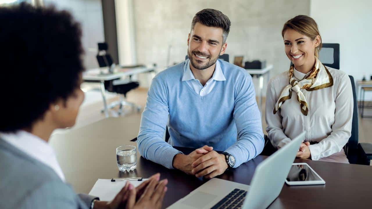 A smiling couple seated at a desk with an interviewer.