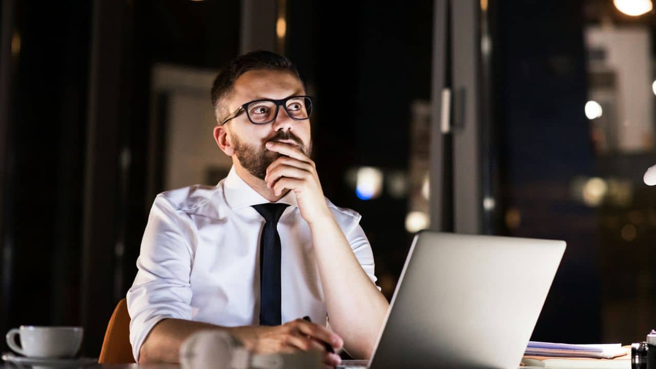 A thoughtful man in a white shirt and tie sits at a desk with a laptop.