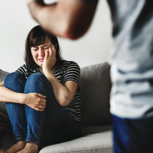 A woman sits crying on a couch, covering her face, while a man's back is visible in the foreground.
