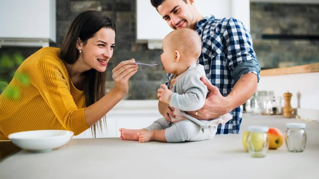 A mother feeding her baby a spoonful of food while the father holds the child on a kitchen counter.
