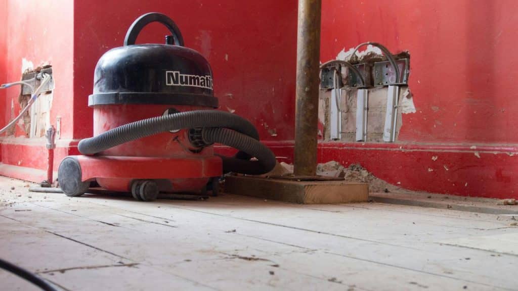 A red Numatic shop vacuum sitting on a dusty wooden floor by a red wall.