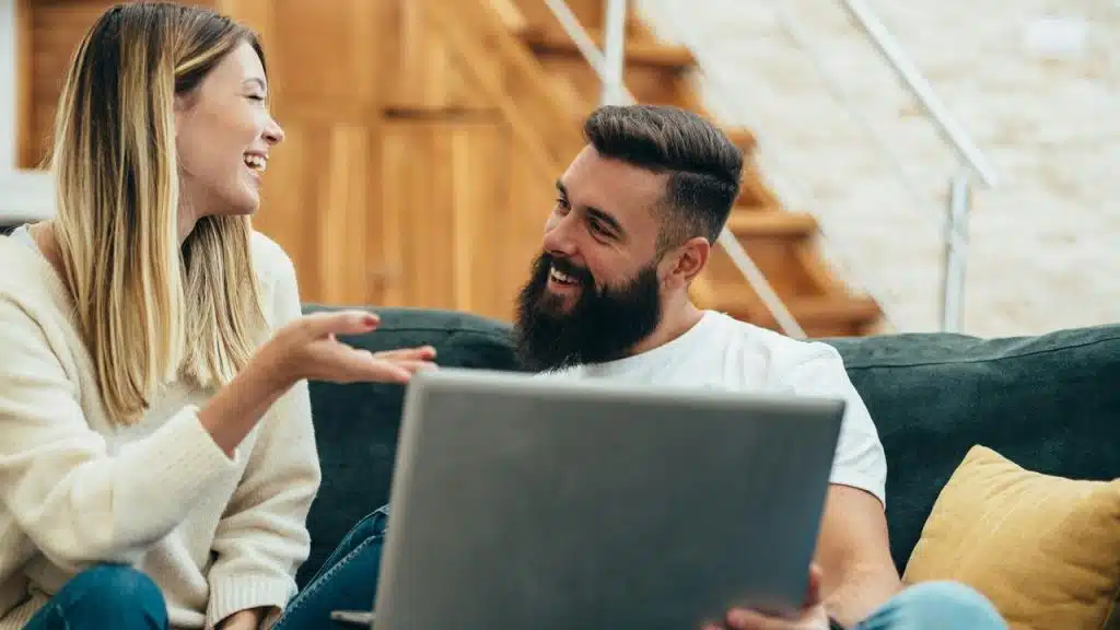A bearded man and woman laughing together on a sofa while using a laptop.