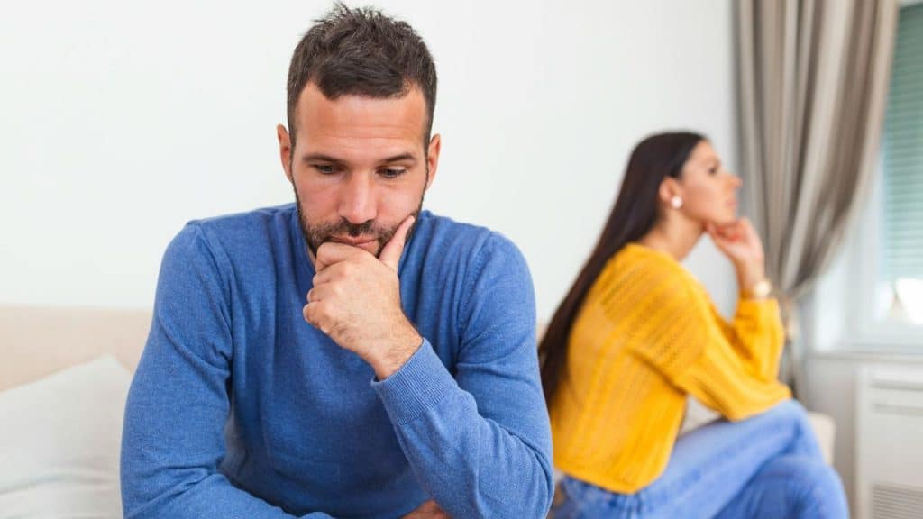 A pensive man in a blue sweater sits with his chin on his hand, while a woman in yellow sits with her back to him in the background.