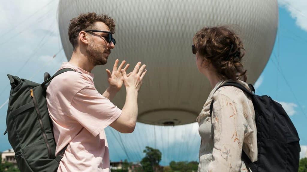 A man gestures expressively while talking to a woman, a large white balloon behind them.