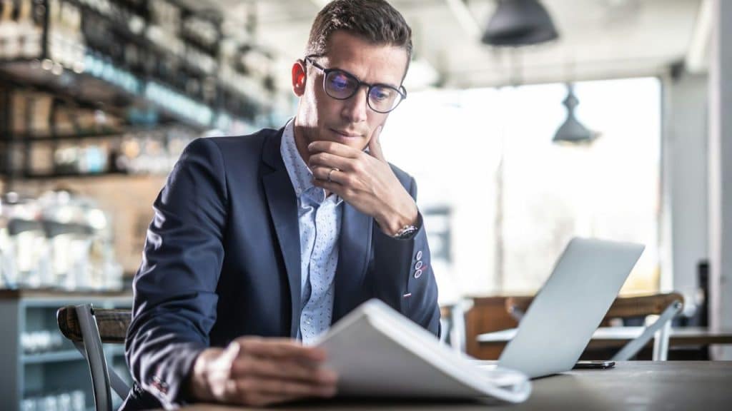 A man in glasses and a suit reads papers at a table with a laptop.