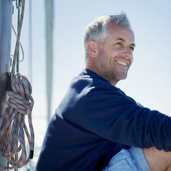 A smiling, grey-haired man in a blue shirt sits on a boat, looking right.