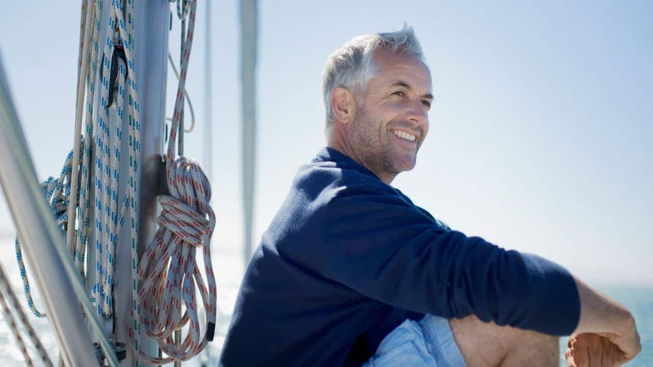A smiling, grey-haired man in a blue shirt sits on a boat, looking right.