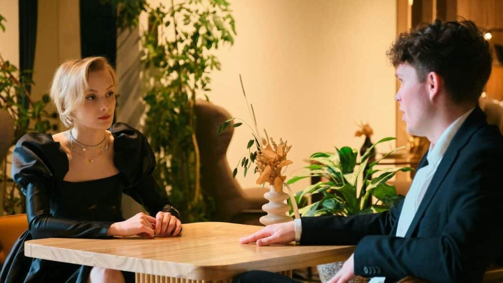 A man in a suit and a woman in a black dress sit opposite each other at a wooden table in a restaurant.