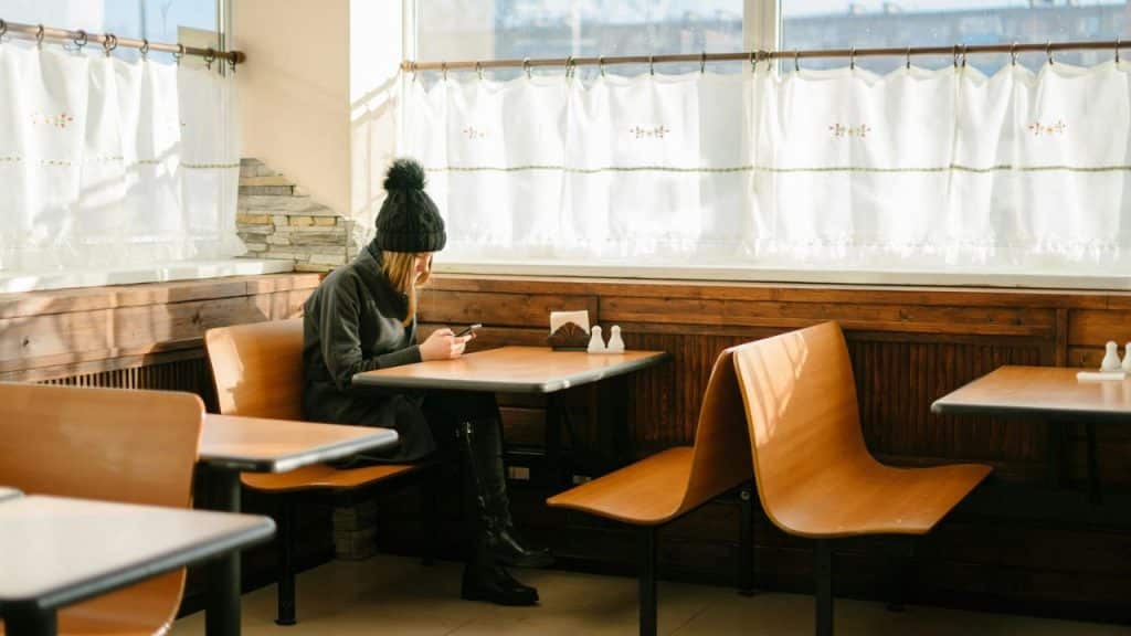 A woman in a dark coat and hat sits alone at a table, looking at her phone.