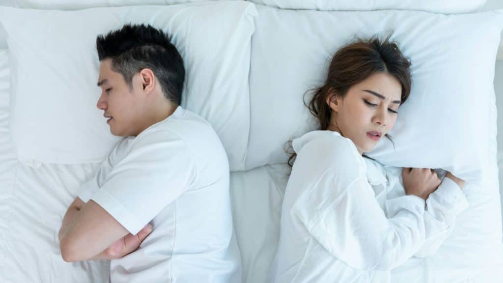 A man and woman lie in bed facing away from each other, looking upset.