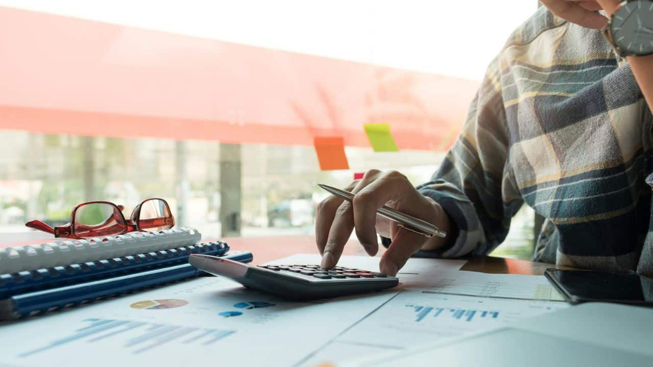 A person in a plaid shirt uses a calculator at a desk with papers and glasses.