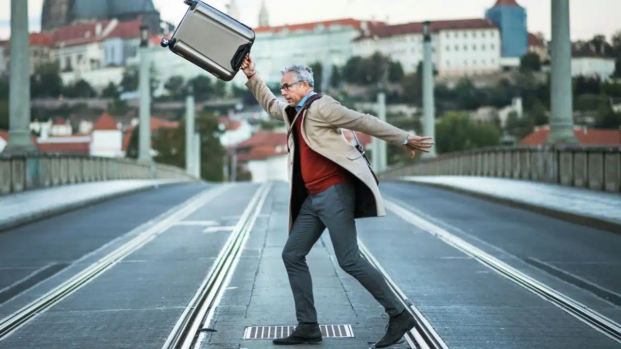 A man in a coat and glasses throws a suitcase in the air on a bridge.