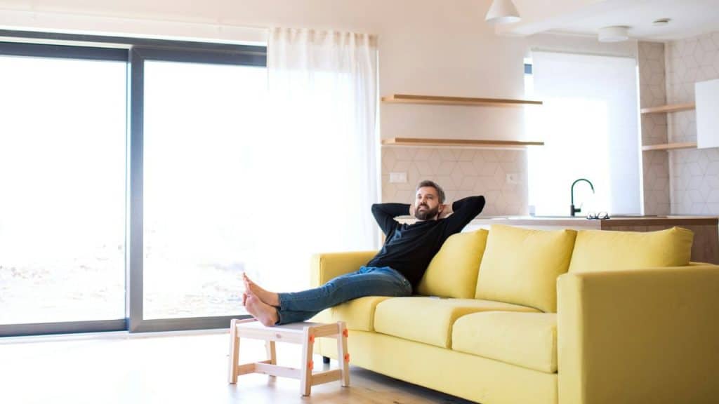 A relaxed man reclines on a yellow sofa in a bright, modern living room.