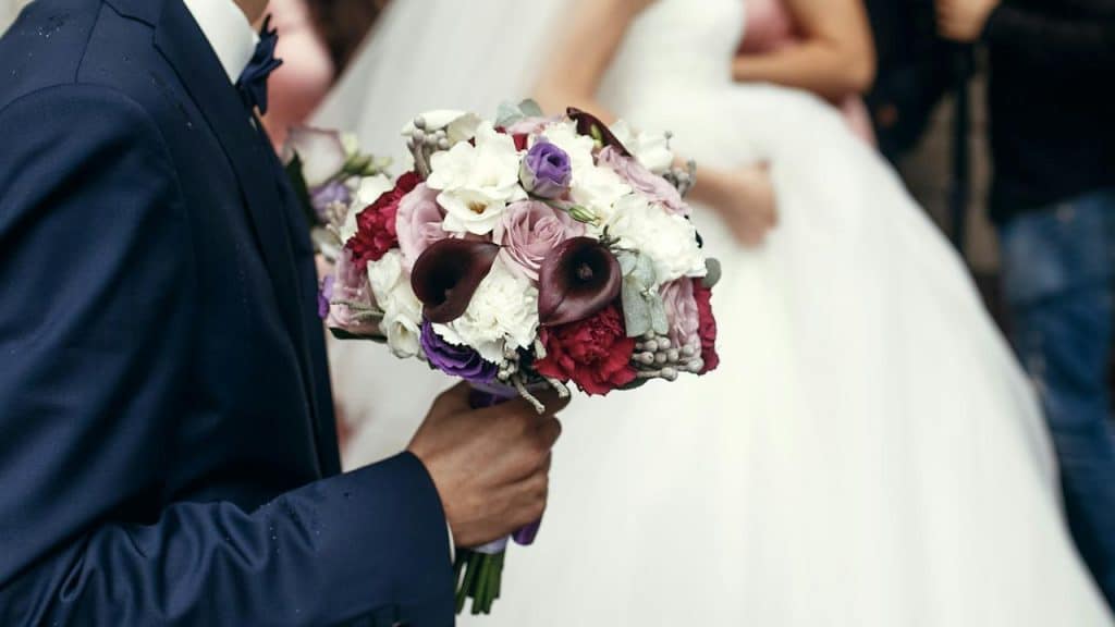 A groom in a navy suit holding a wedding bouquet of white, red, and purple flowers.