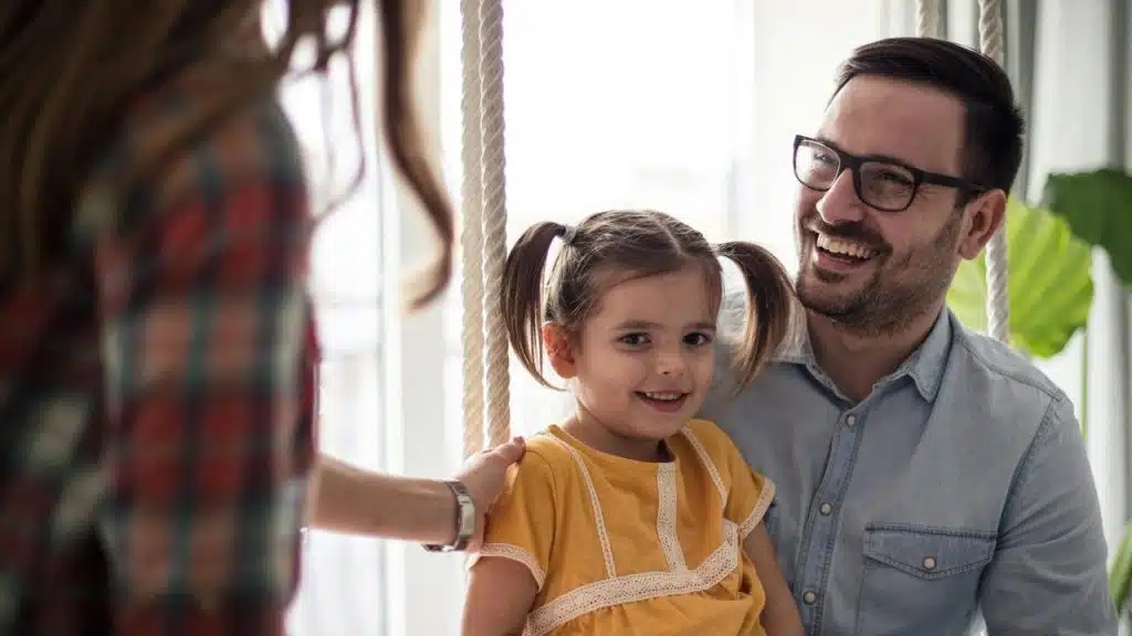 A smiling father holds his young daughter on an indoor swing as a woman steadies it.