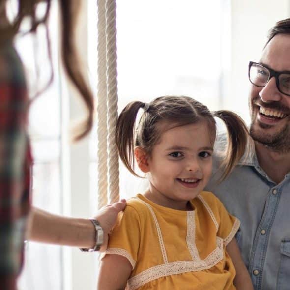 A smiling father holds his young daughter on an indoor swing as a woman steadies it.