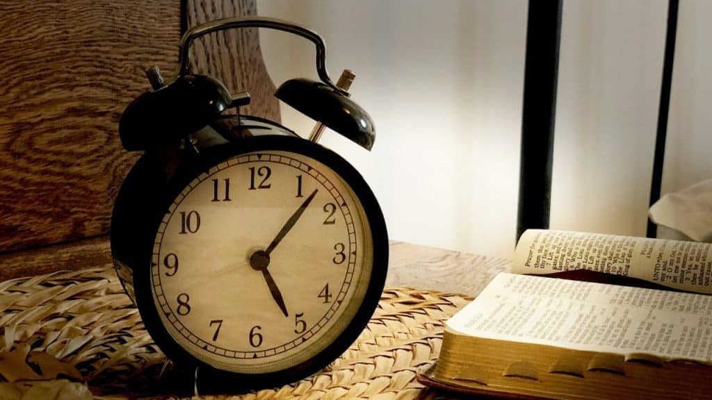 A classic black alarm clock sits on a woven mat beside an open book.
