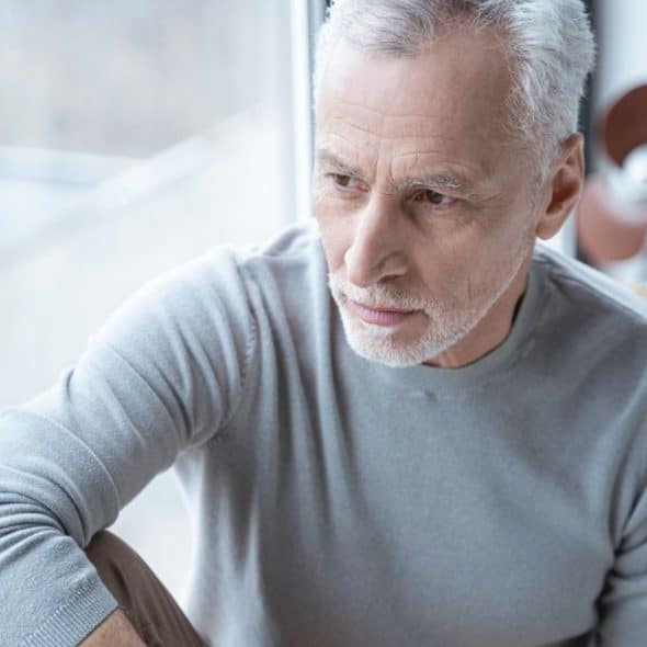 A serious senior man with grey hair and a beard looks out a window.