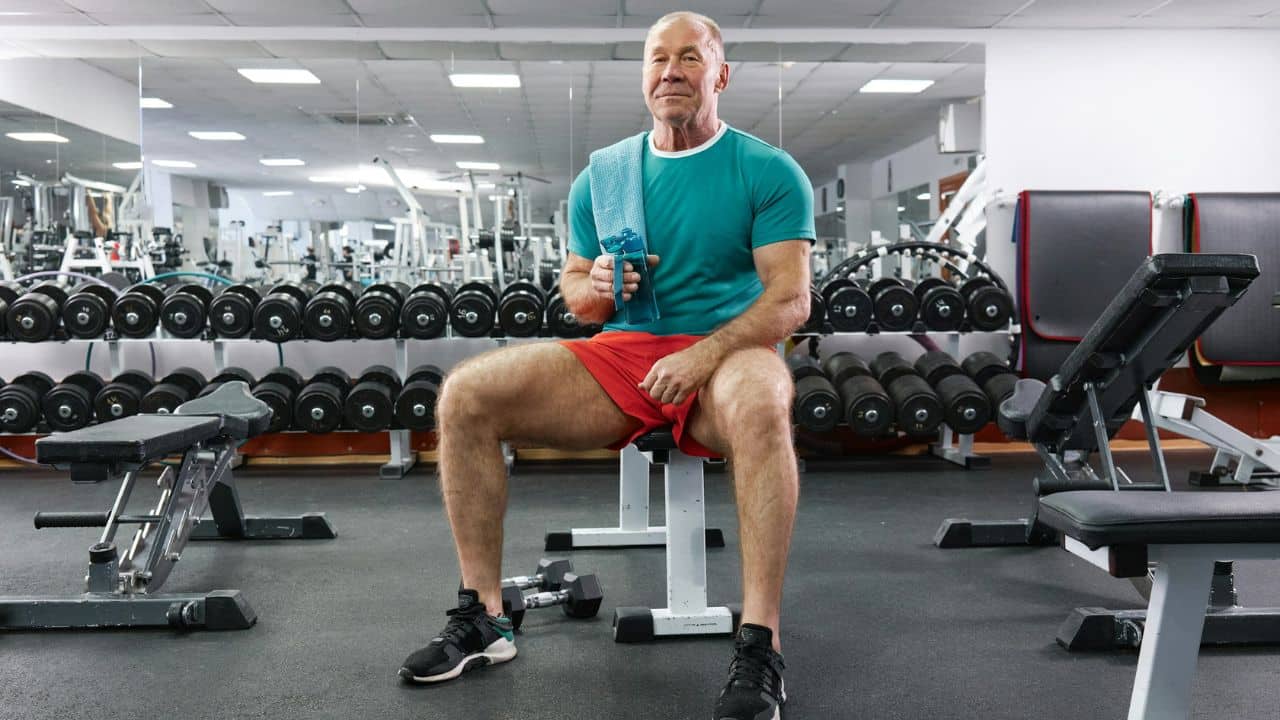 A fit, smiling man in a teal shirt and red shorts sits on a gym bench.