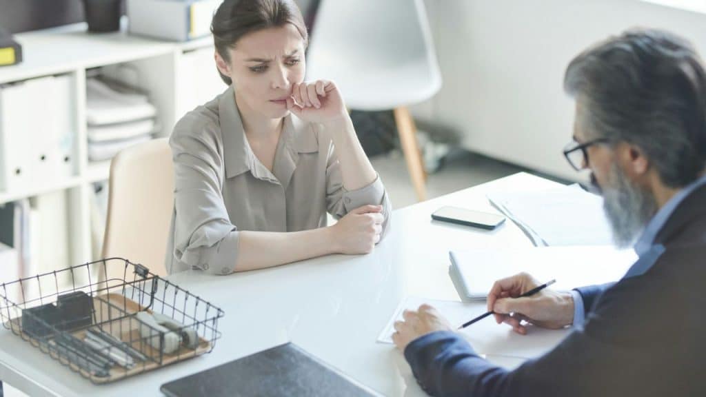 A concerned woman sits across a desk from a man writing on paper during an interview.
