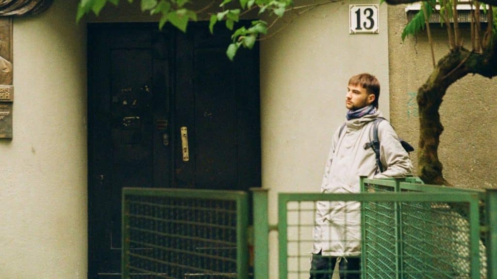 A young man with a backpack stands outside a building with the number 13.