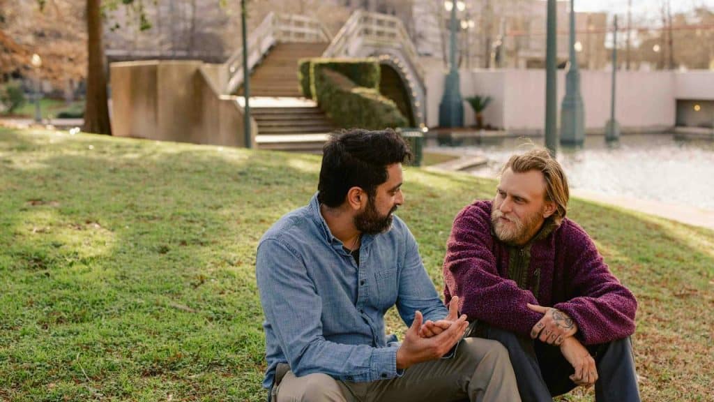 A pair of men sitting on grass talking by a pond.