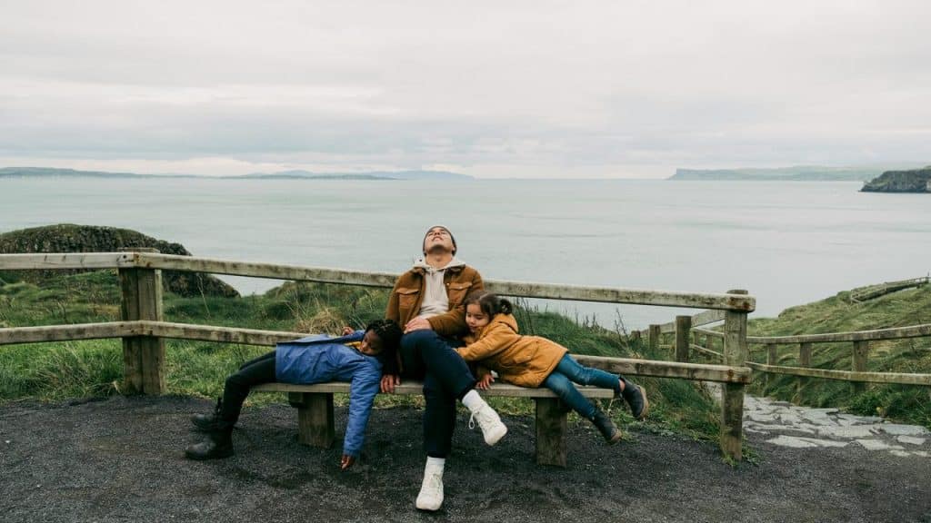 A parent dozing on a seaside bench with two children draped beside them.