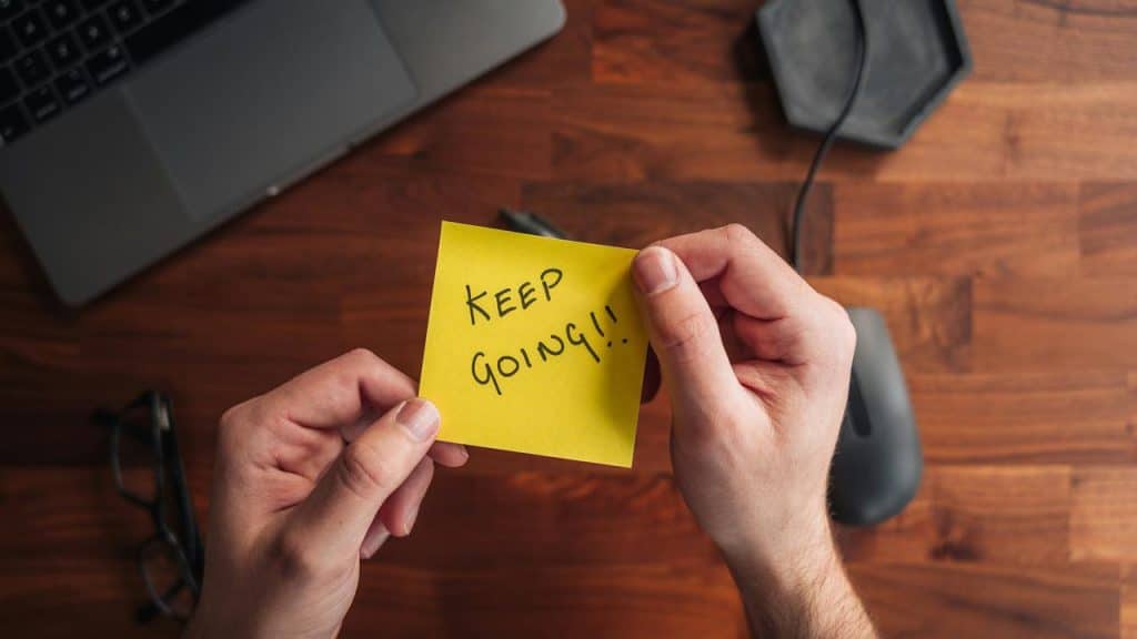 A pair of hands holding a yellow sticky note that reads “KEEP GOING!!”.