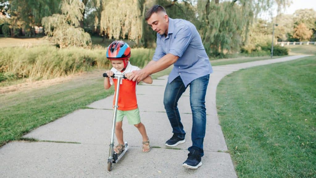 A man guiding a young boy on a scooter along a paved park path.