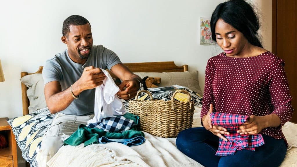A smiling couple is sitting on a bed, folding laundry together from a basket.