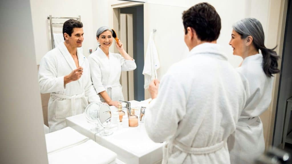 A couple in bathrobes smiles at each other in the mirror while brushing hair and teeth.