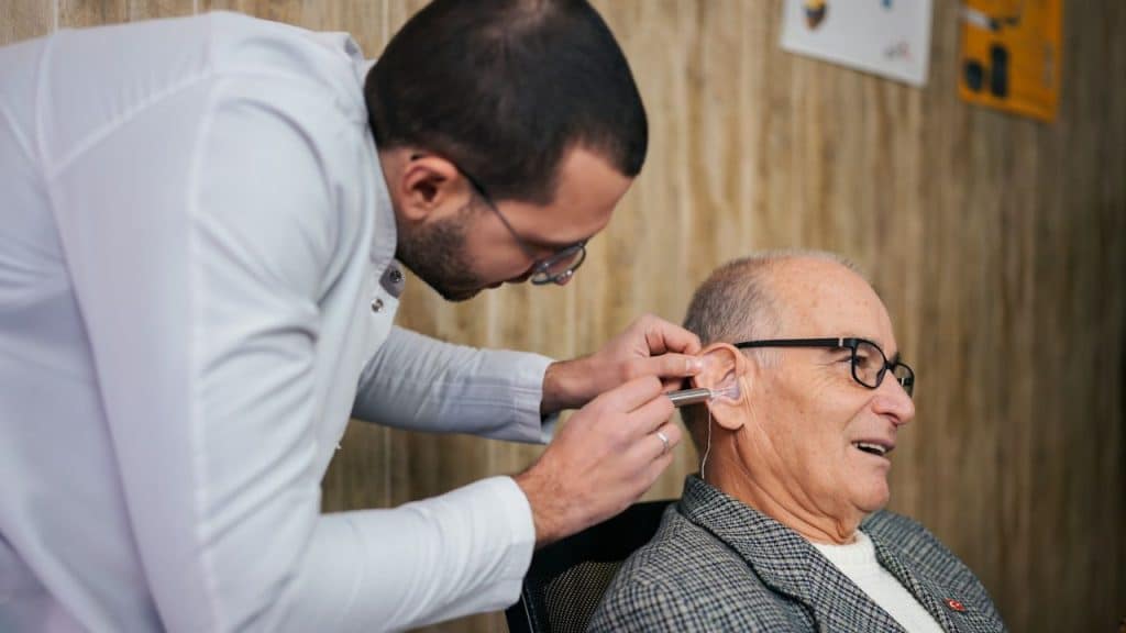 A man in a white coat adjusts a device in an elderly man's ear.