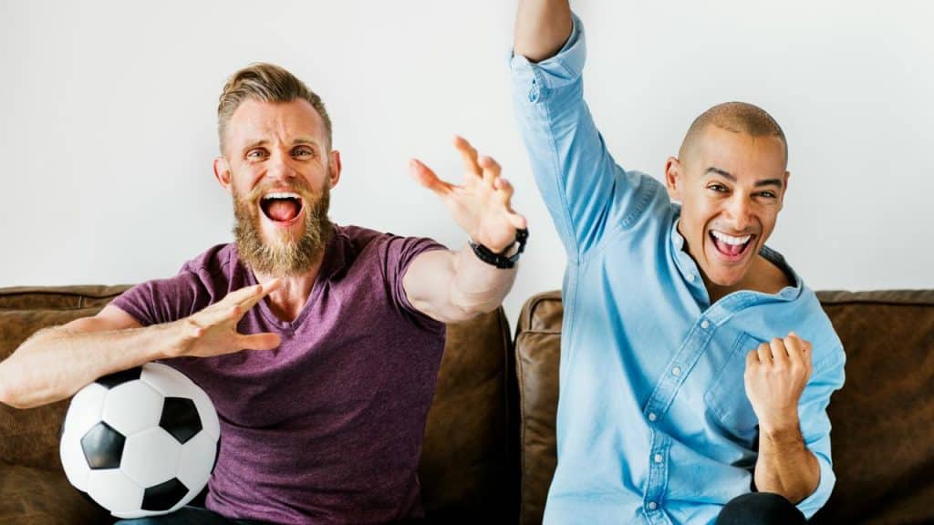 A man and his friend cheering on a couch, one holding a soccer ball.