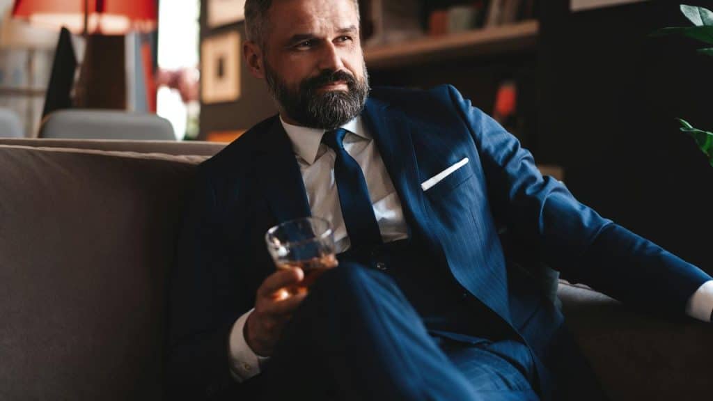 A man in a blue suit sits on a sofa, holding a glass of whiskey.