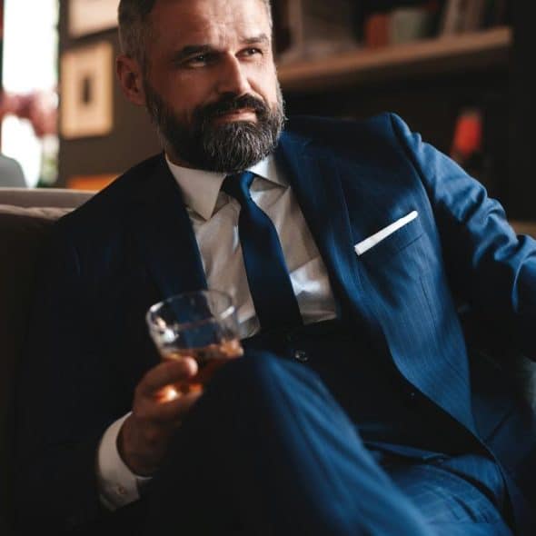 A man in a blue suit sits on a sofa, holding a glass of whiskey.