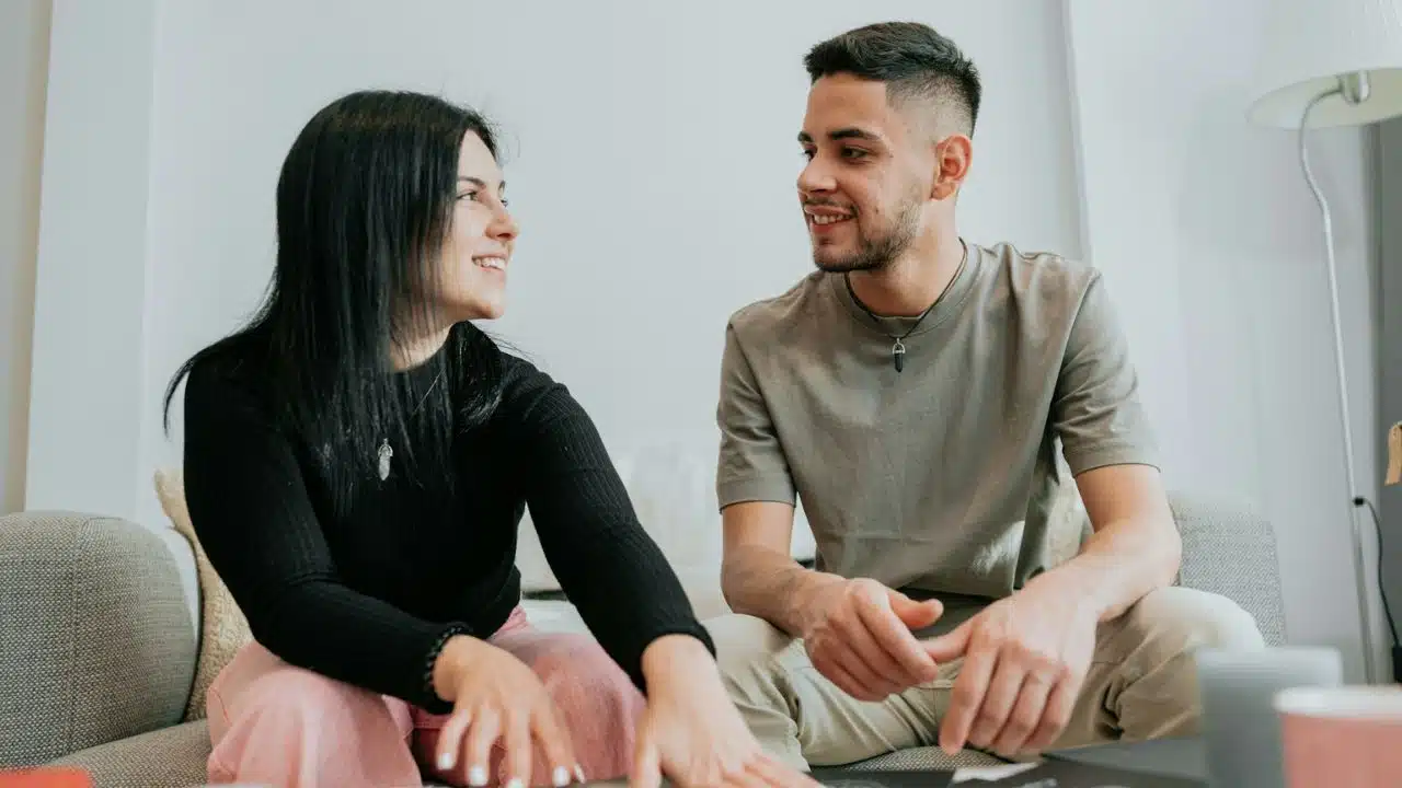 A woman with black hair smiles at a man on a couch.