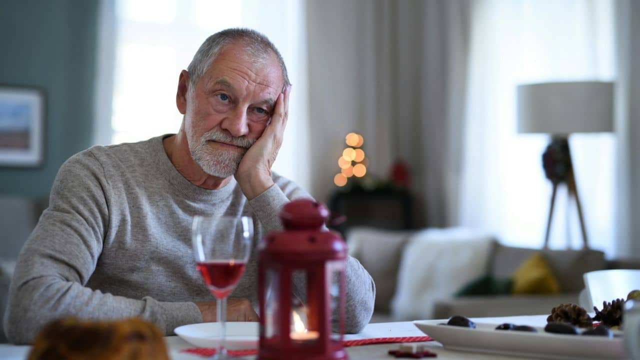 A sad, gray-haired man rests his head on his hand at a table with wine.