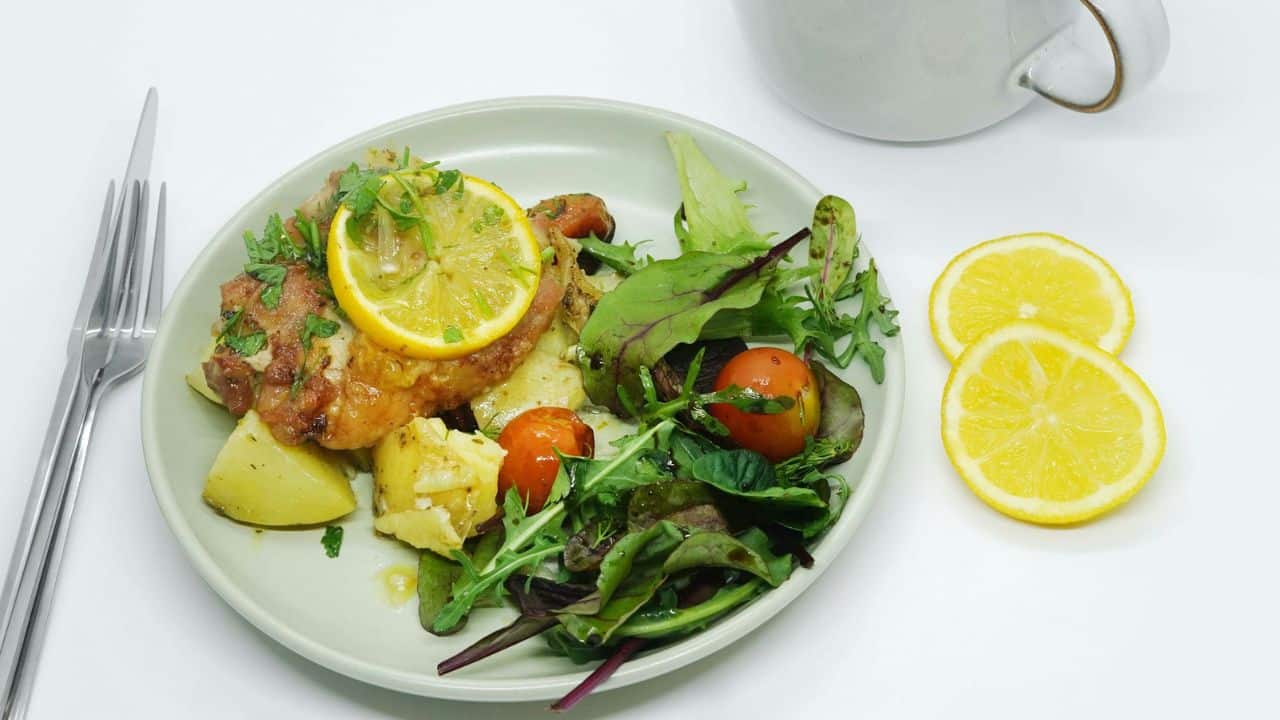A plate of roasted chicken, potatoes, salad, and lemon slices sits on a white surface.