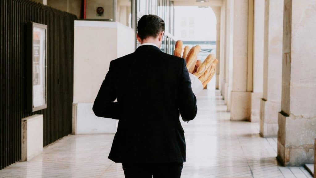 A man in a suit walks away from the camera, holding several baguettes.