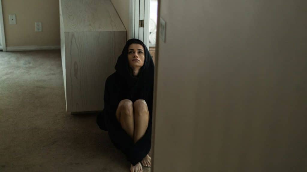 A young woman in a black hoodie sits on a carpeted floor, looking up.