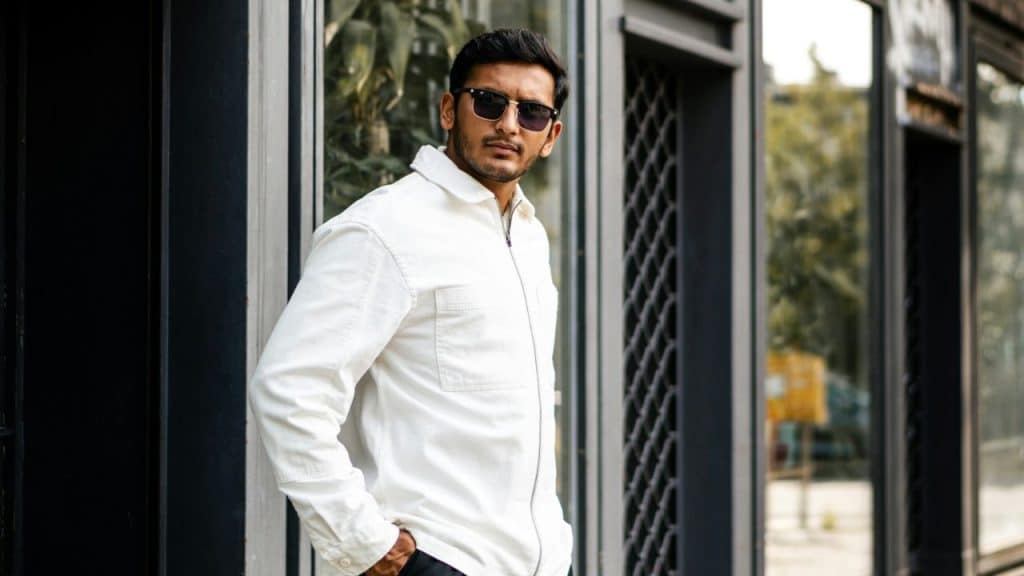 A stylish man in a white jacket and sunglasses poses confidently on a city street.