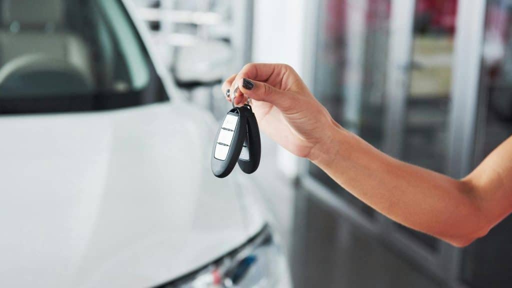A hand holding car key fobs in front of a car.